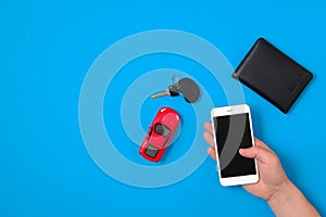 Car rental app concept. Toy car, car key, auto drive license, human hand holding smartphone on blue background. Flat lay