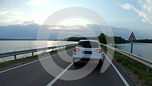 Car riding through road near lake. White SUV driving at dam route on summer day. Follow to auto moving through bridge of