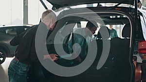 Car salesman showing customer car in automotive dealership showroom. Details in the open trunk of the car. Smiling