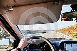 Car salon, steering wheel, hand of woman on it, mirror and view on landscape with road and nature at sunny summer or autumn day.