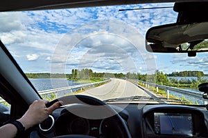 Car salon, steering wheel, hand of woman and view on nature landscape. Road, forest, blue sky, white clouds at sunny day. Concept