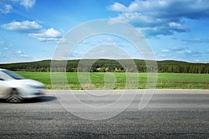 The car speeds along a country road, surrounded by forest and blue sky