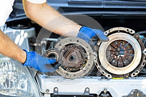 Car technician holding used clutch assembly - friction, pressure plate and throwout bearing in front of the vehicle engine