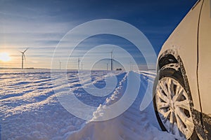 Car tires on winter road covered with snow. Vehicle on snowy country road with wind turbine in the background. Snow covered field