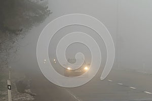 Car traveling on the foggy road with headlights or headlamps on. Low visibility - Dangerous driving of cars in winter in bad