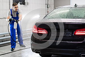 Car with turned on backlights standing in car wash service. Worker cover the car with foam with high-pressure sprayer.