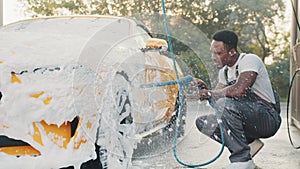 Car wash outdoors concept. Luxury modern car in foam. Young African man with high pressure jet spraying the foam on car