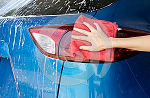 Car wash service. Car covered with white soap foam. Man hand holding red microfiber cloth and polish tail light of blue car. Auto