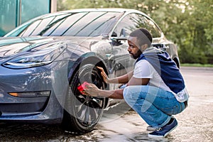 Car wash service outdoors. Car wash self-service concept. Handsome African guy wiping wheel of his modern luxury