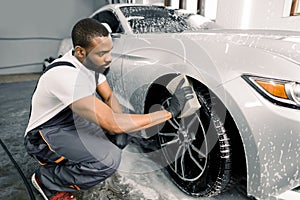 Car washing and detailing photo. African man worker in protective overalls and rubber gloves, washing car wheel rims of