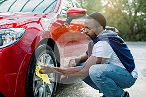 Car washing on open air. Young hipster African bearded man cleaning a wheel, car rims of modern luxury red car with