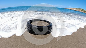 A car wheel in beach flooded with waves and sand, a rubber wheel in sea with sea foam, concept of ocean and sea pollution, polluti