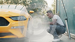 Car wheel, rim or alloy, wash outdoors. Car cleaning with water jet. Horizontal shot of pleasant African guy in white t