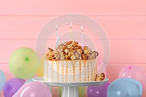 Caramel drip cake decorated with popcorn and pretzels near balloons against pink wooden background