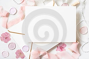 Card and envelope near pink decorations, seals and silk ribbons on white table top view, wedding mockup