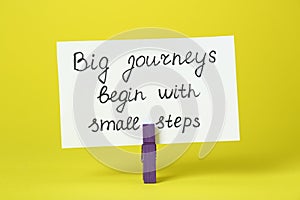 Card with phrase Big Journeys Begin With Small Steps and clothespin on yellow background. Motivational quote