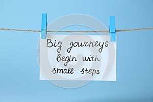 Card with phrase Big Journeys Begin With Small Steps hanging on rope against light blue background. Motivational quote