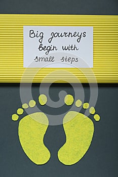 Card with phrase Big Journeys Begin With Small Steps and paper feet on dark background, top view. Motivational quote