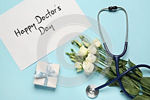 Card with phrase Happy Doctor\'s Day, stethoscope, gift box and eustoma flowers on light blue background, flat lay