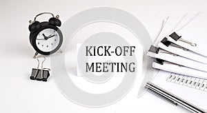 Card with text KICK OFF MEETING on a white background, near office supplies and alarm clock. Business concept