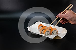 A cardboard box for sushi, a man hand using chopsticks takes a roll. Black background. Place for text. Asian cuisine concept.