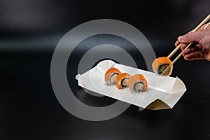 A cardboard box for sushi, a man hand using chopsticks takes a roll. Black background. Place for text. Asian cuisine concept.