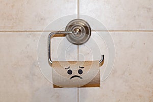 Cardboard center of a toilet paper roll without paper with and sad face drawn hanging hanging from a toilet paper roll holder