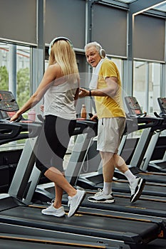 Cardio training to fat burning. Smiling senior man with headphones talks to woman on treadmill in fitness center.