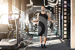 Cardio workout. Confident muscular man jumping with skipping rope in gym interior