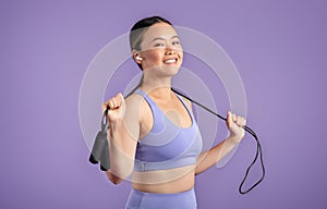 Cardio workout. Smiling asian woman in sportswear holding skipping rope around neck, posing on purple background