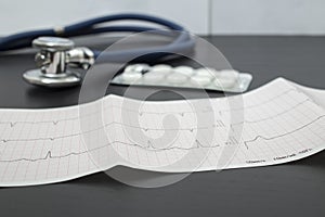 A cardiogram, a blue stethoscope and pills are lying on a dark table in a doctor`s office, against the background of a white wall
