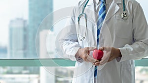 Cardiologist doctor holding red heart with professional medical care in clinic office with hospital city building background