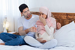 Care and support concept. Arab expecting wife feeling sick, husband giving her glass of water, sitting on bed