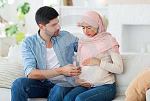 Care and support during pregnancy. Muslim expecting woman feeling sick, man giving her glass of water, siting on sofa