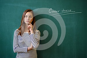 Career choice concept. Young female high school student standing in front of chalkboard in classroom, looking at camera.