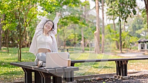 A carefree Asian woman sits on a bench in a green park, enjoying nature while working remotely