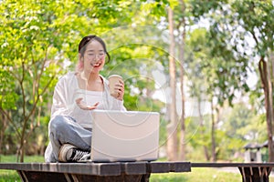 A carefree Asian woman sits on a bench in a green park, using her laptop, having an online meeting