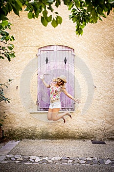 Carefree beautiful woman in hat jumping midair in front of old wooden closed window of textured building wall. Ecstatic hipster