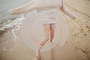 Carefree beautiful woman in knitted sweater running on sandy beach at cold sea waves, having fun. Stylish young happy female