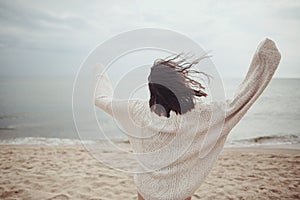 Carefree beautiful woman in knitted sweater and with windy hair running on sandy beach at cold sea, having fun. Stylish young