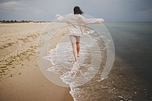 Carefree beautiful woman in knitted sweater and with windy hair running on sandy beach at cold sea, having fun. Stylish young