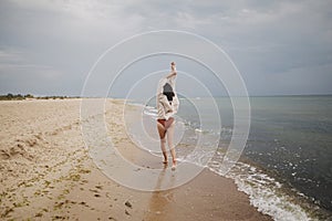 Carefree beautiful woman in knitted sweater and with windy hair running on sandy beach at cold sea, having fun. Stylish young sexy