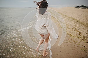 Carefree beautiful woman in knitted sweater and with windy hair running on sandy beach at cold sea, having fun. Stylish young sexy