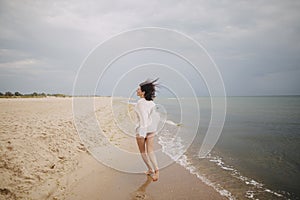 Carefree beautiful woman in knitted sweater and with windy hair running on sandy beach at cold sea waves, having fun. Stylish