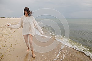 Carefree beautiful woman in knitted sweater and with windy hair running on sandy beach at cold sea waves, having fun. Stylish