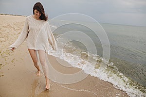 Carefree beautiful woman in knitted sweater and with windy hair running on sandy beach at cold sea waves, having fun. Stylish