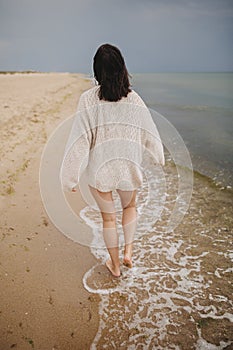 Carefree beautiful woman in knitted sweater and with windy hair walking on sandy beach at cold sea. Stylish young sexy female