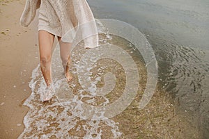 Carefree beautiful woman running barefoot on sandy beach with cold sea waves, having fun. Cropped view. Stylish happy female