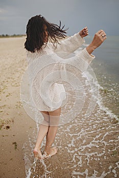 Carefree beautiful woman with windy hair running on sandy beach at cold sea waves, having fun. Stylish young happy female in