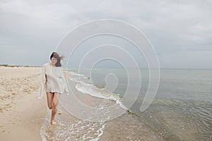 Carefree beautiful woman with windy hair running on sandy beach at cold sea waves, having fun. Stylish young happy female in